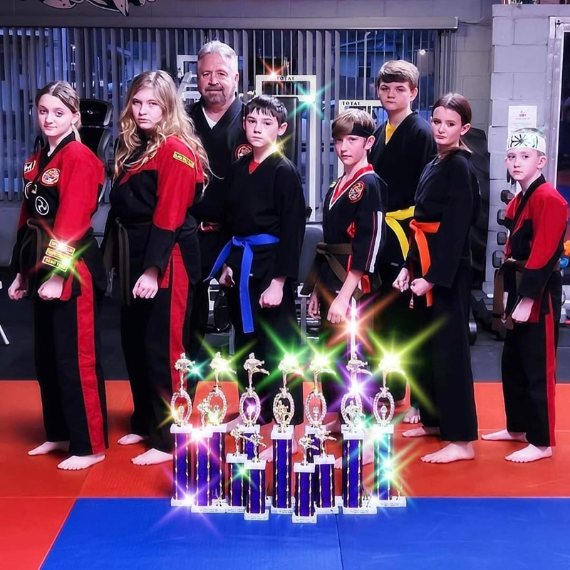 A group of martial artists in uniforms pose behind a row of trophies on a mat in a training gym.