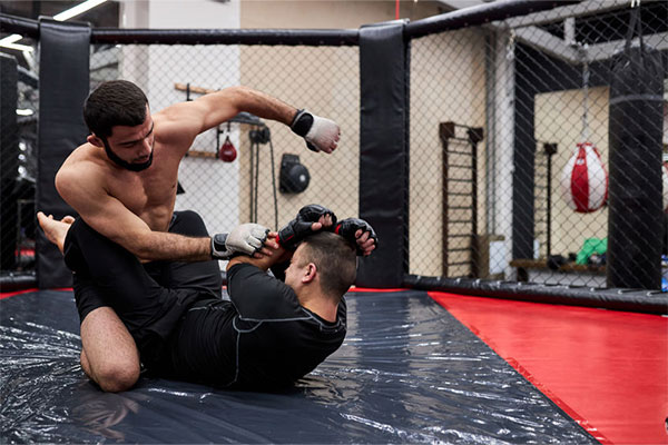 Two men practicing mixed martial arts sparring in a gym, one delivering a punch from a mounted position.